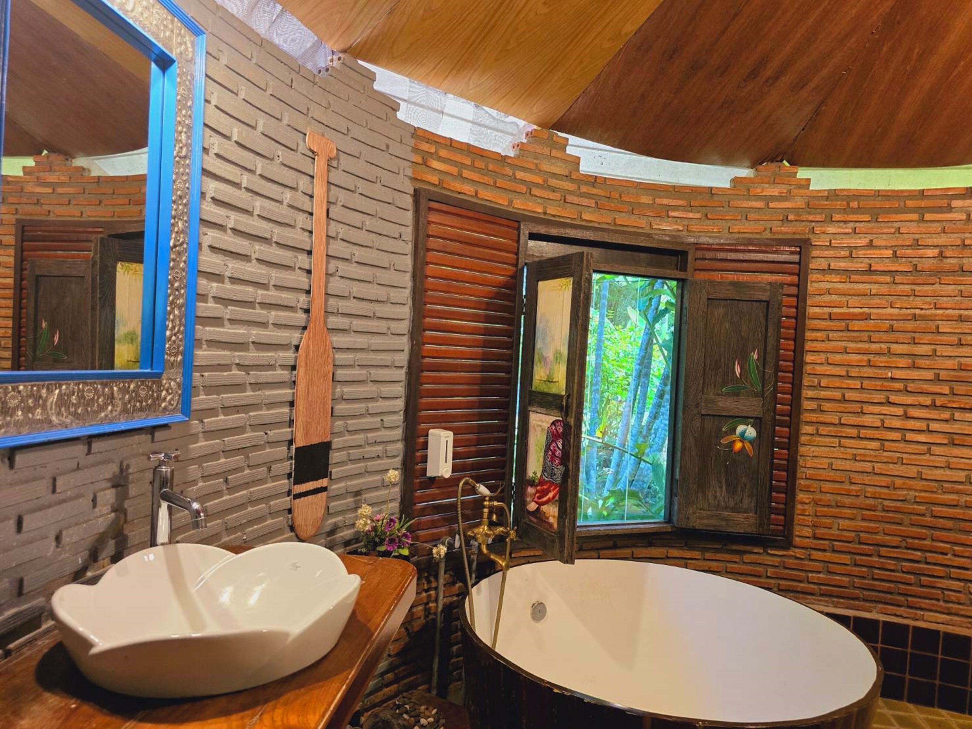 A bathroom with a wooden vanity, a large oval bathtub, a mirror with a blue frame, and a window with a view of greenery.