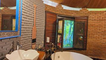 A bathroom with a wooden vanity, a large oval bathtub, a mirror with a blue frame, and a window with a view of greenery.