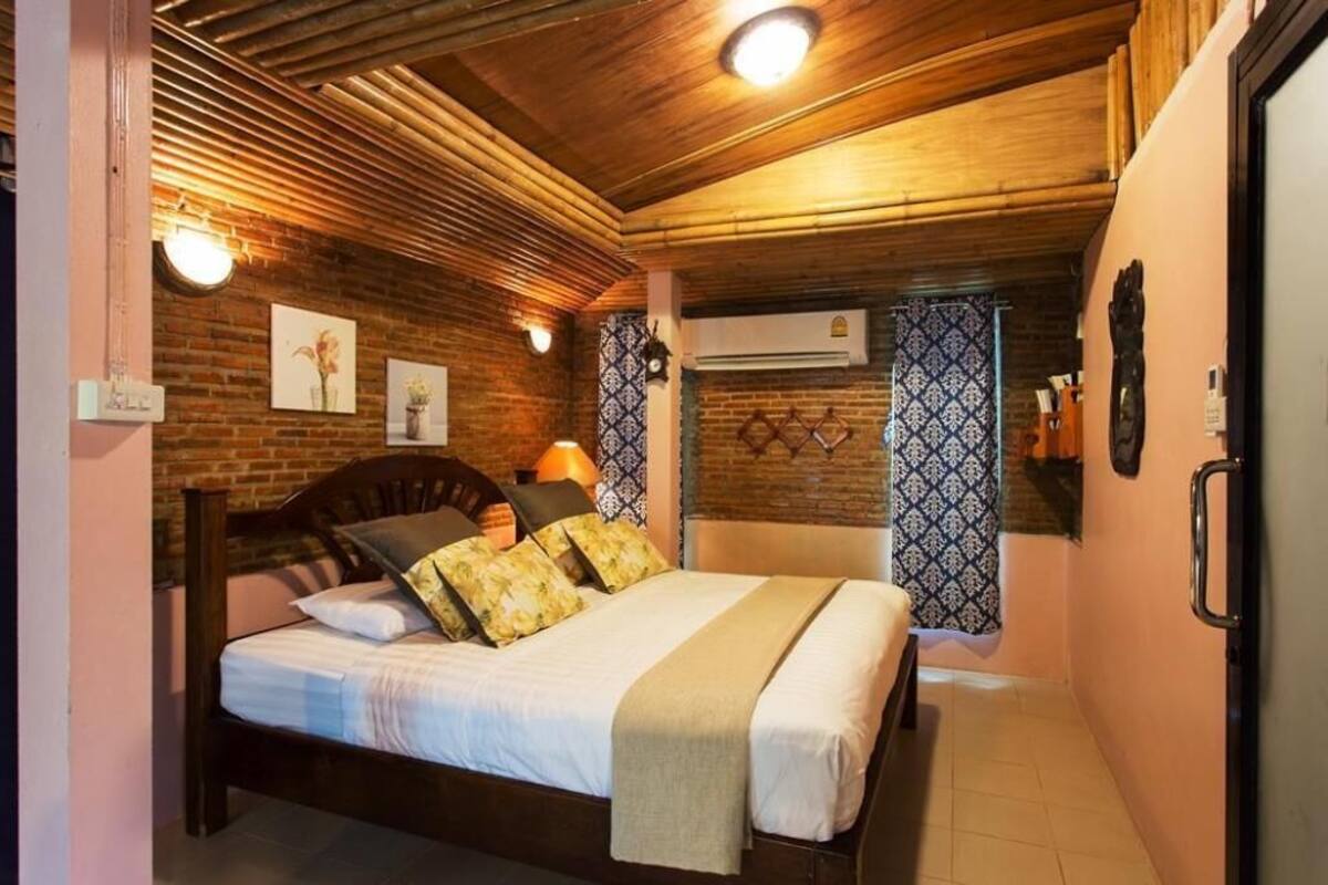 Un dormitorio con techo de madera, una cama con muchas almohadas, una lámpara de noche y una cortina estampada.