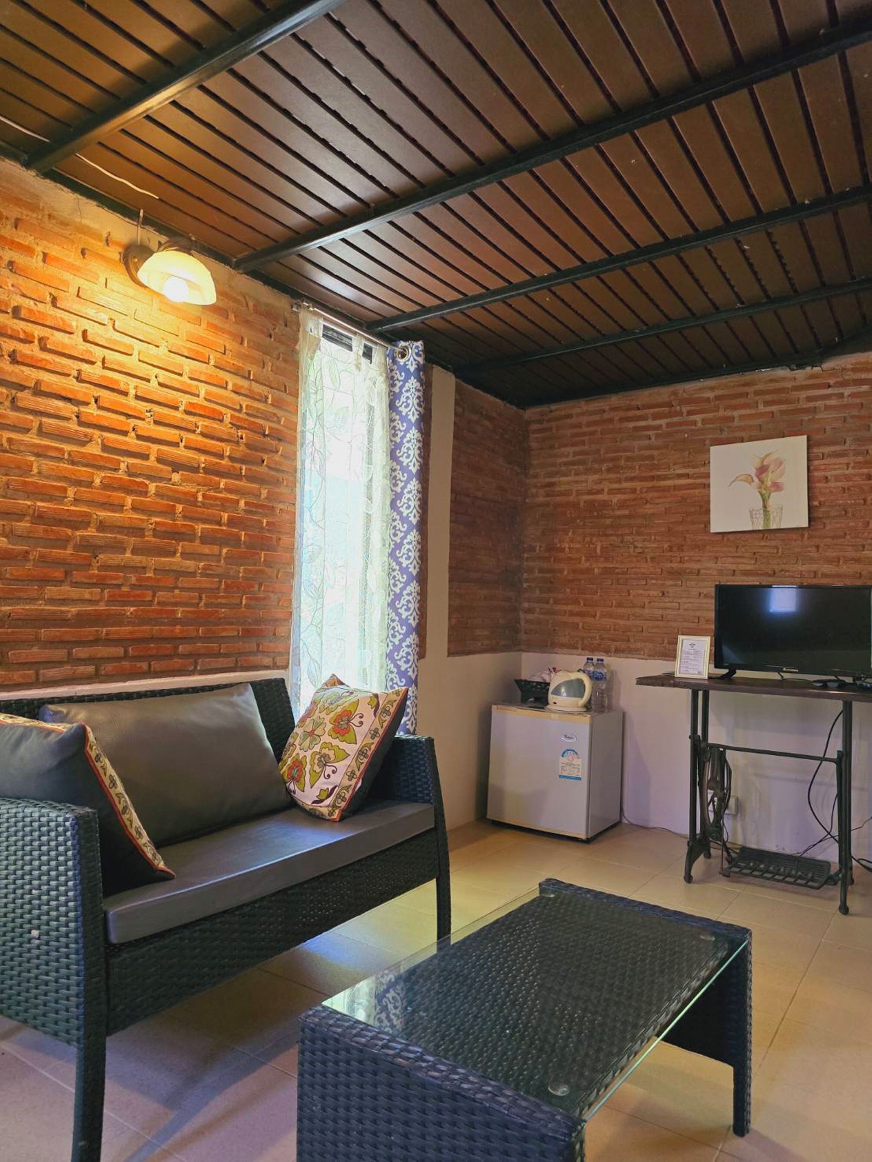 Une pièce confortable avec un mur en briques, un canapé avec des coussins, une petite table, un téléviseur et un réfrigérateur.
