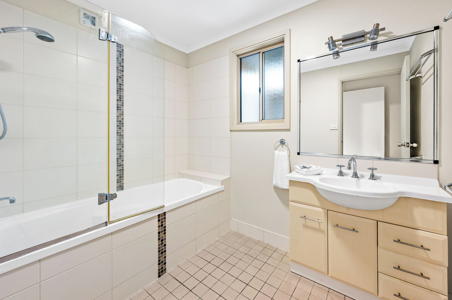 Un baño moderno con ducha de vidrio, una bañera blanca, un lavabo con espejo y muebles de color beige.