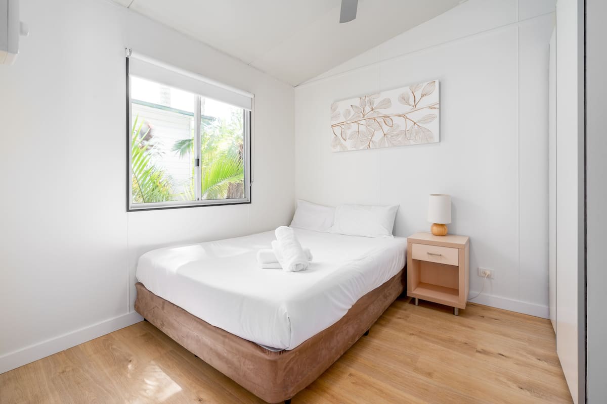 A bedroom with a bed, a nightstand, a wall art piece, and a window with a view of greenery.