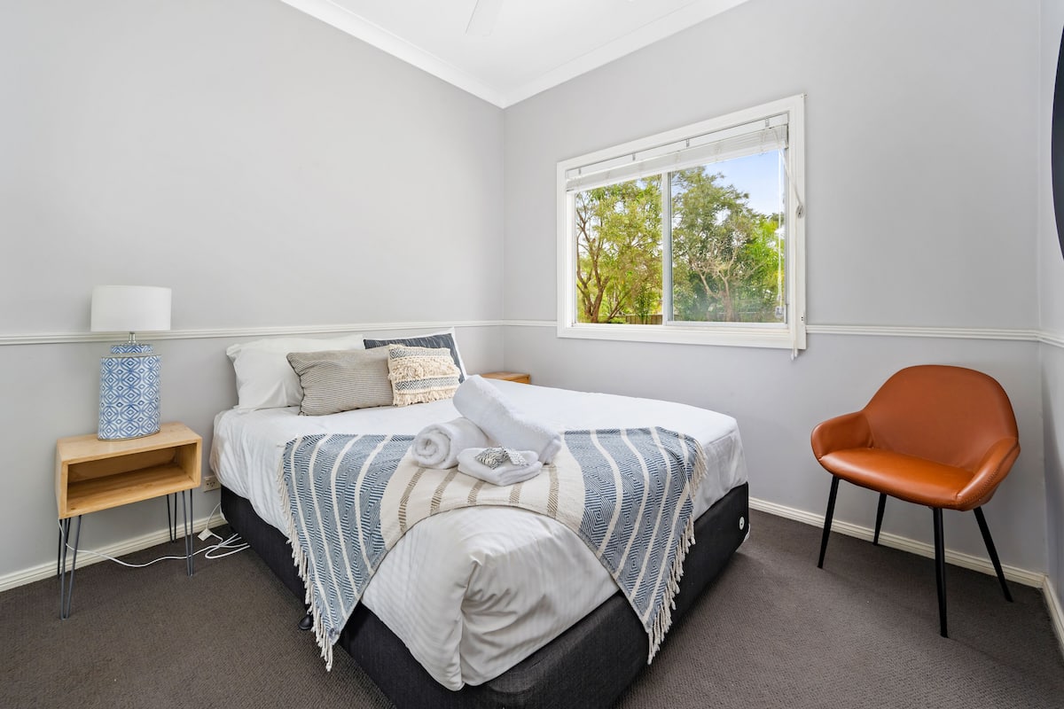 A bedroom with a bed, a chair, a nightstand, and a window with a view of trees.