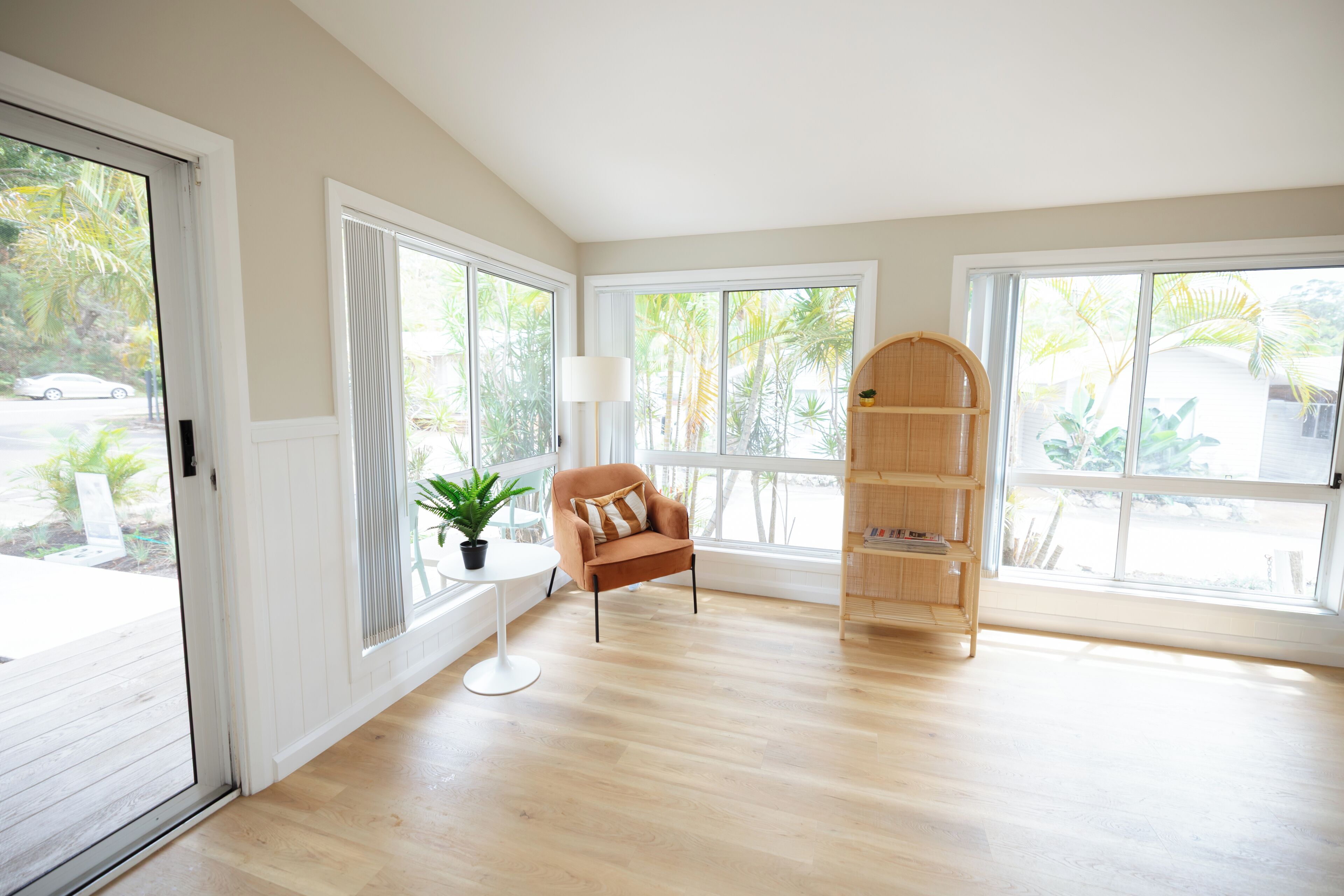 A spacious room with large windows, a wooden floor, a brown armchair, a small table with a plant, and a wooden bookshelf.