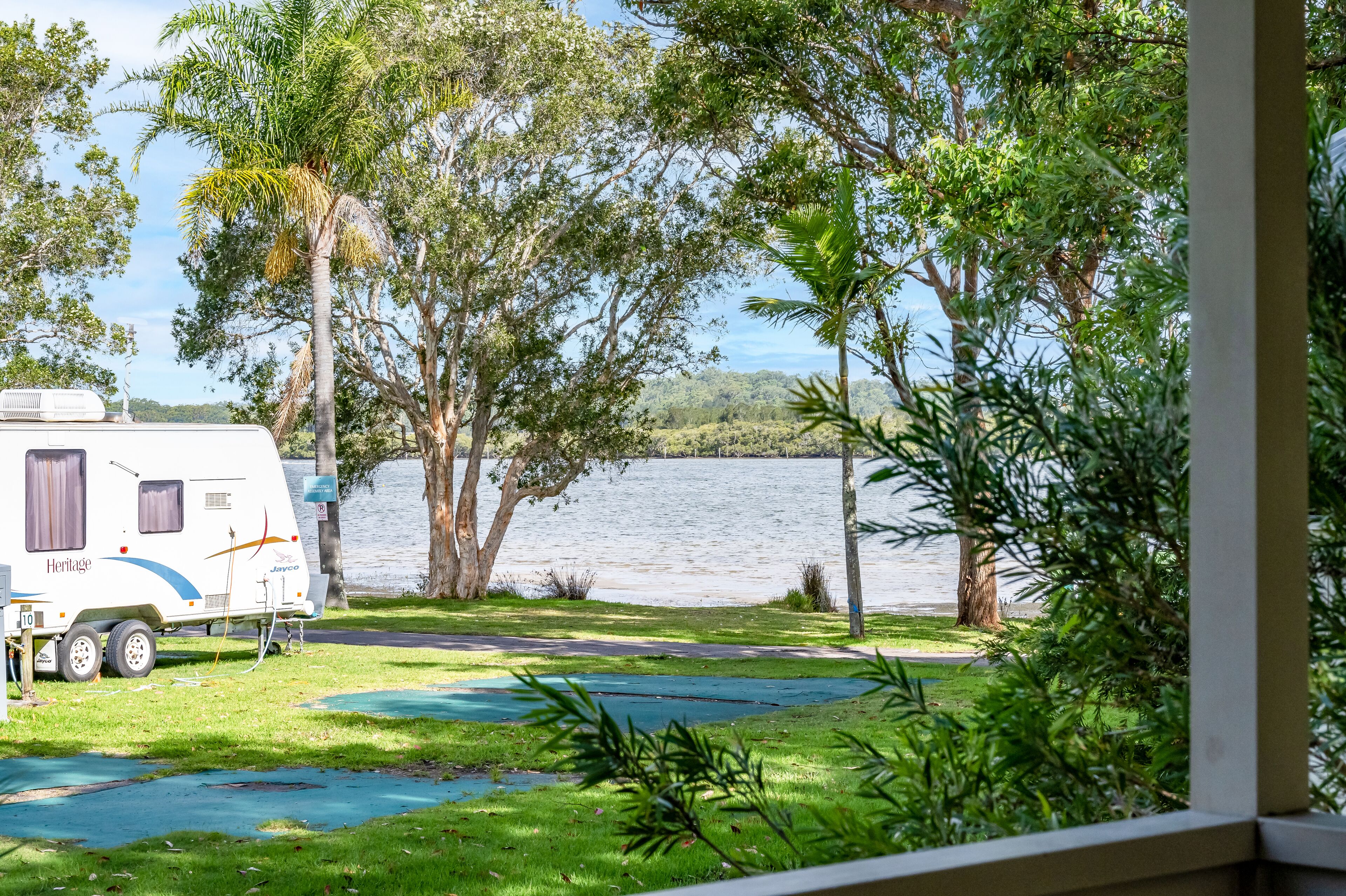 Una caravana blanca llamada 'Heritage' estacionada cerca de un lago con árboles y una zona verde.