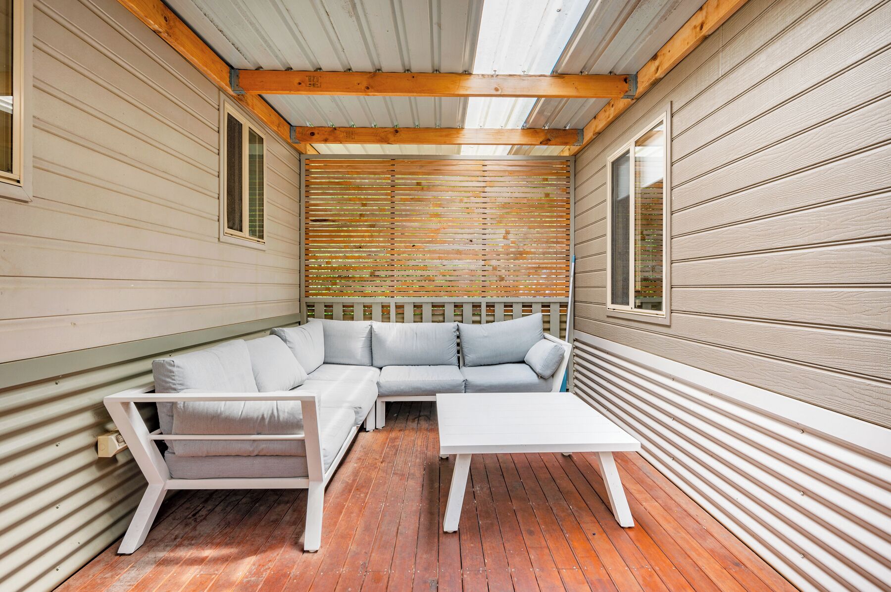 Un espace extérieur aménagé en coin salon, de dimensions réduites, avec un canapé blanc et une petite table, le tout entouré de murs en bois et d’une clôture en bois à lattes.