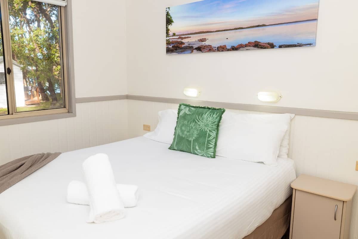 A neatly made bed with white linens and a green pillow, a window with a view of trees and a building, and a painting of a beach scene on the wall.