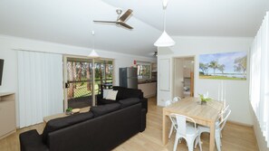 A modern living room with a black sofa, a wooden dining table with white chairs, a ceiling fan, and a view of a garden through a glass door. - Koala Shores Port Stephens Holiday Park (Lemon Tree Passage)