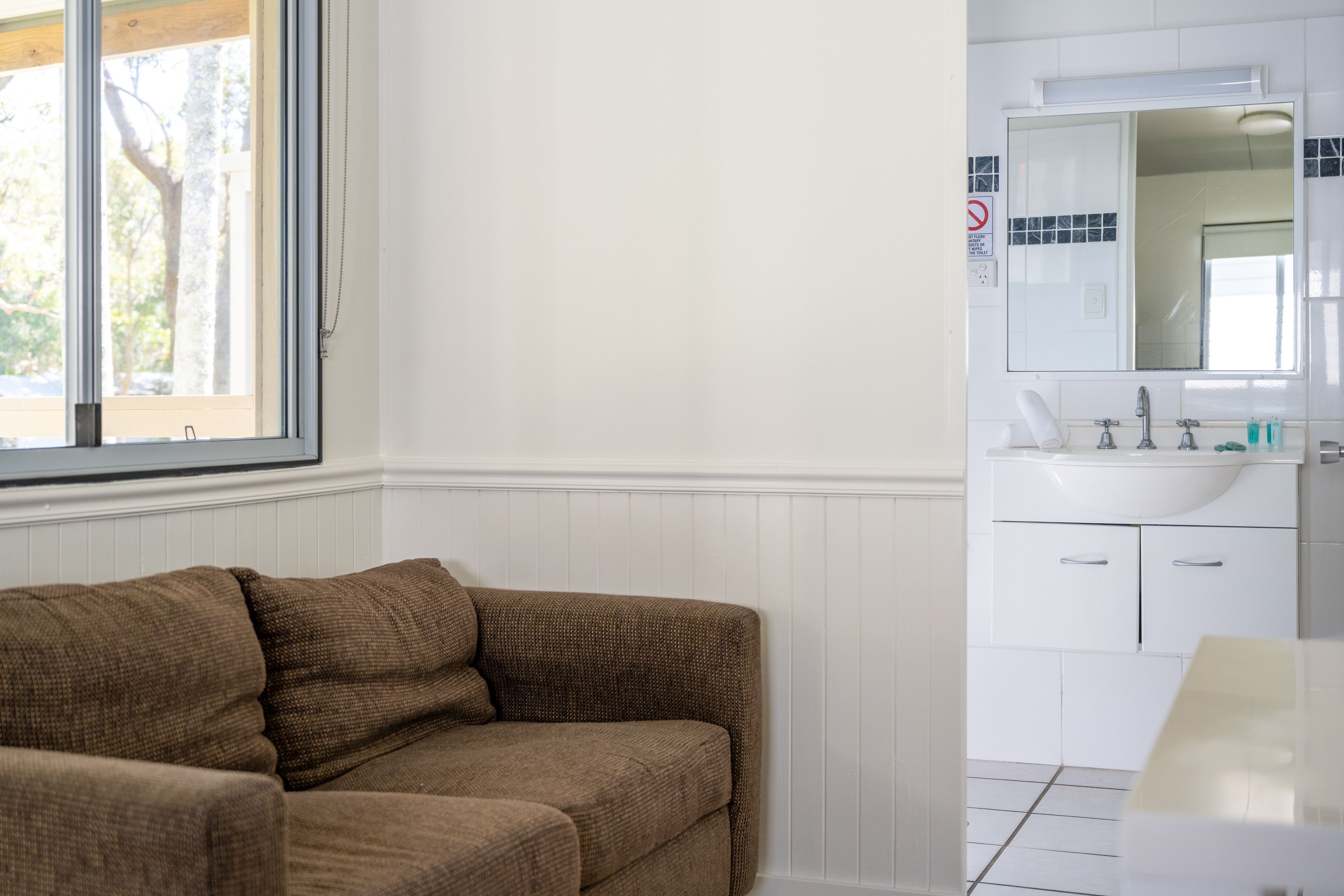 Une pièce avec un canapé marron, un lavabo blanc et une fenêtre donnant sur des arbres.