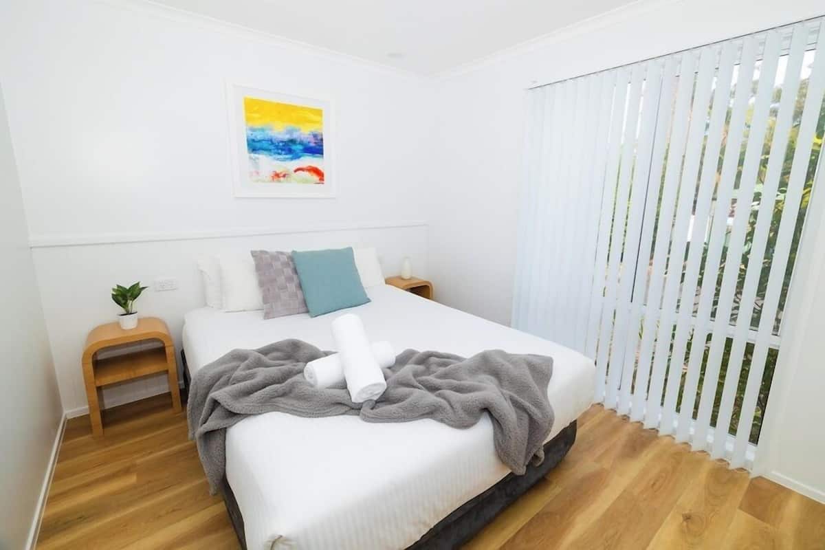 A bedroom with a bed, a bedside table, a nightstand, a window with blinds, and a painting on the wall.