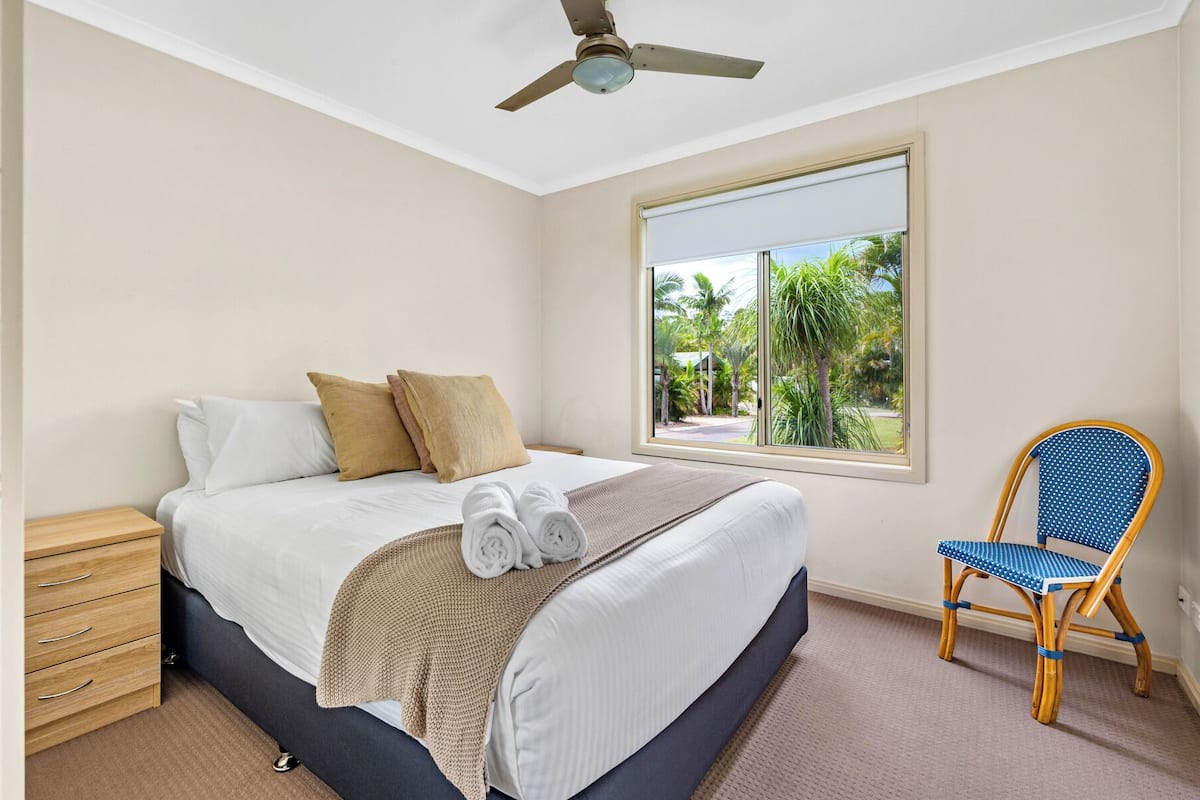 A bedroom with a bed, a wooden nightstand, a chair, and a window with a view of palm trees.