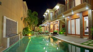 A swimming pool with lounge chairs and a wooden deck, surrounded by buildings with balconies and plants. - O Villas (Keroboken Kelod)
