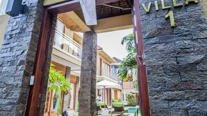 A stone facade with "VILLA 1" signage, a wooden door frame, and a view of a pool area with lounge chairs and umbrellas. - O Villas (Keroboken Kelod)