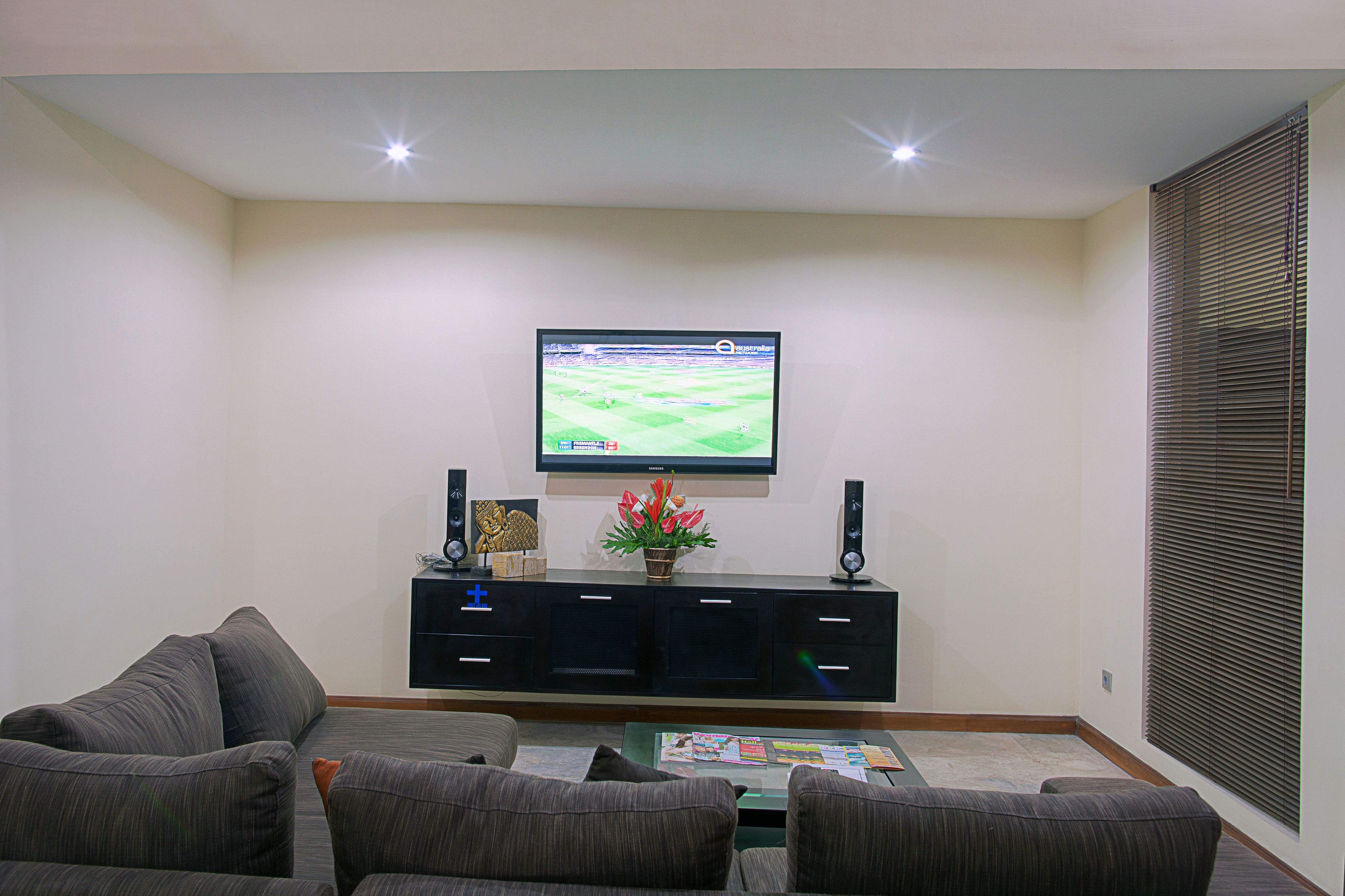 A living room with a flat-screen TV mounted on the wall, a dark-colored entertainment unit below, and a sofa set.