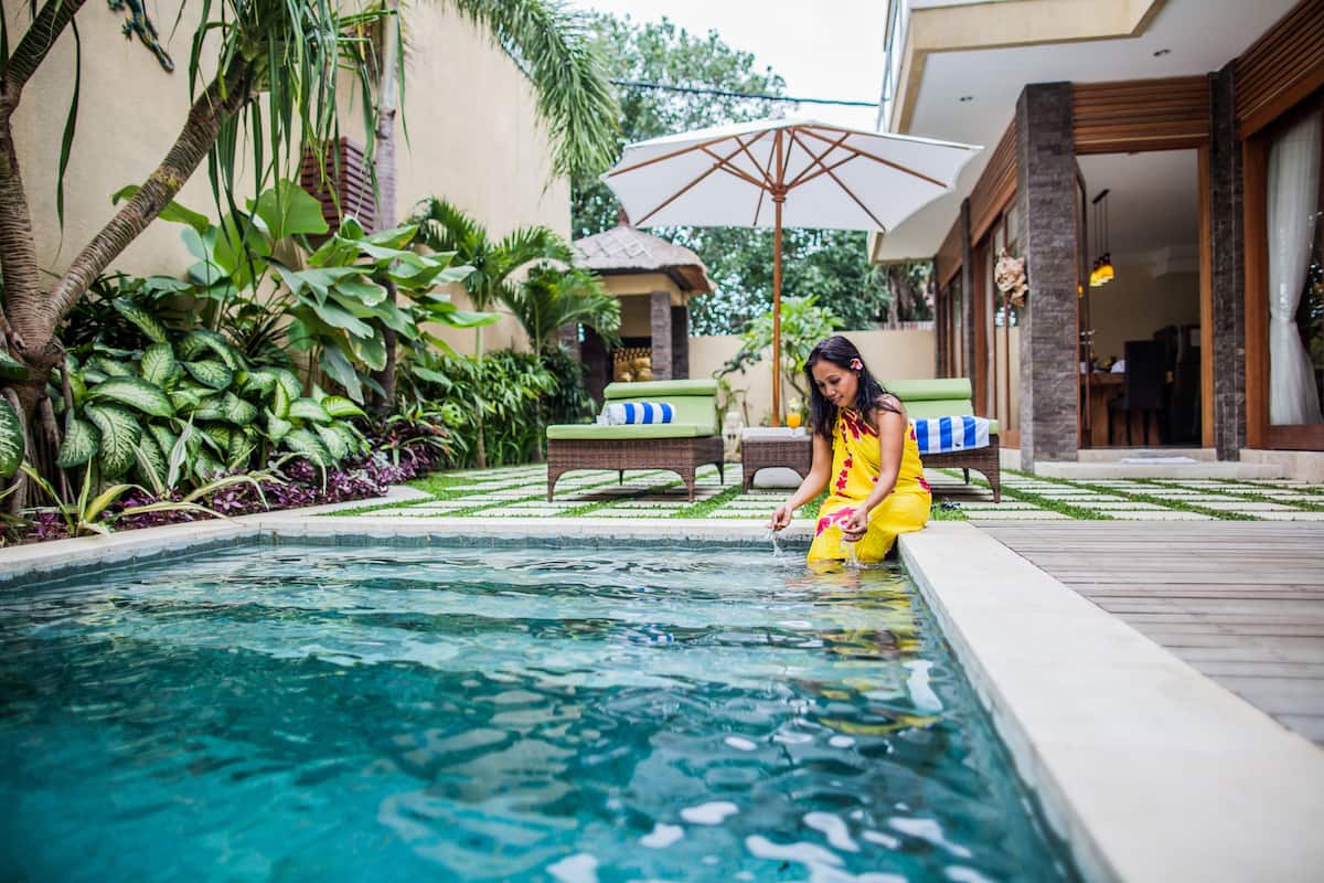 Área de piscina com uma pessoa vestindo um vestido amarelo, cercada por vegetação exuberante e mobiliário de exterior.