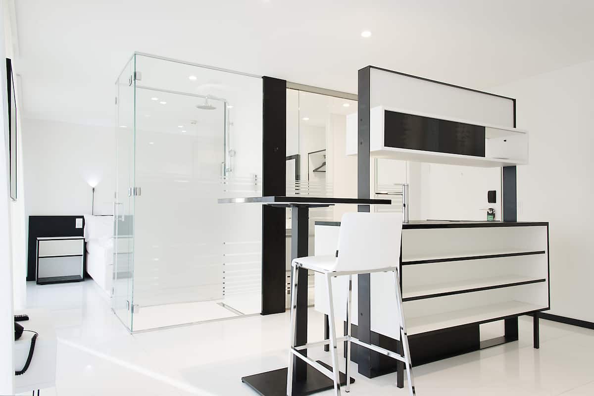 A modern hotel room with a glass shower enclosure, a white desk with a chair, and a black and white console table.