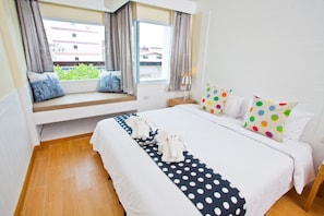 A hotel room with a large bed, a window seat with cushions, a bedside table with a lamp, and a view of buildings outside. - Paradise Hotel (Udon Thani)