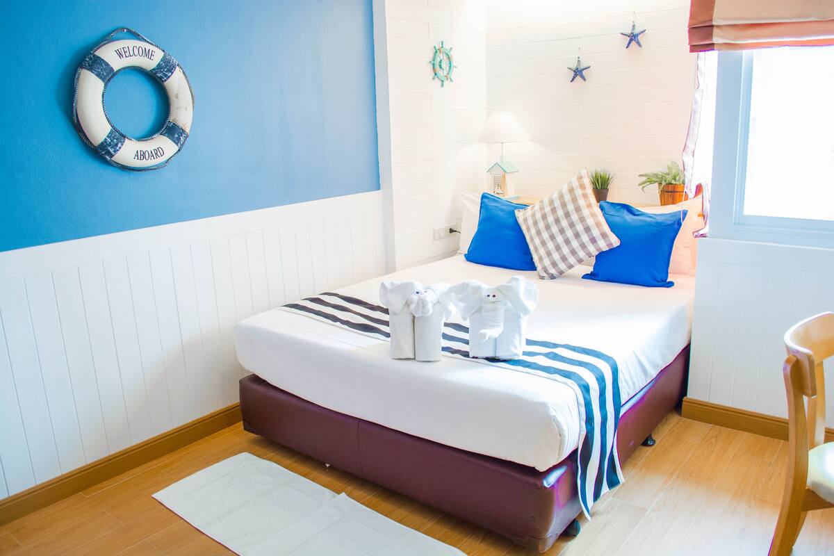 A neatly made bed with a striped blanket, a life ring on the wall, and a window with curtains.