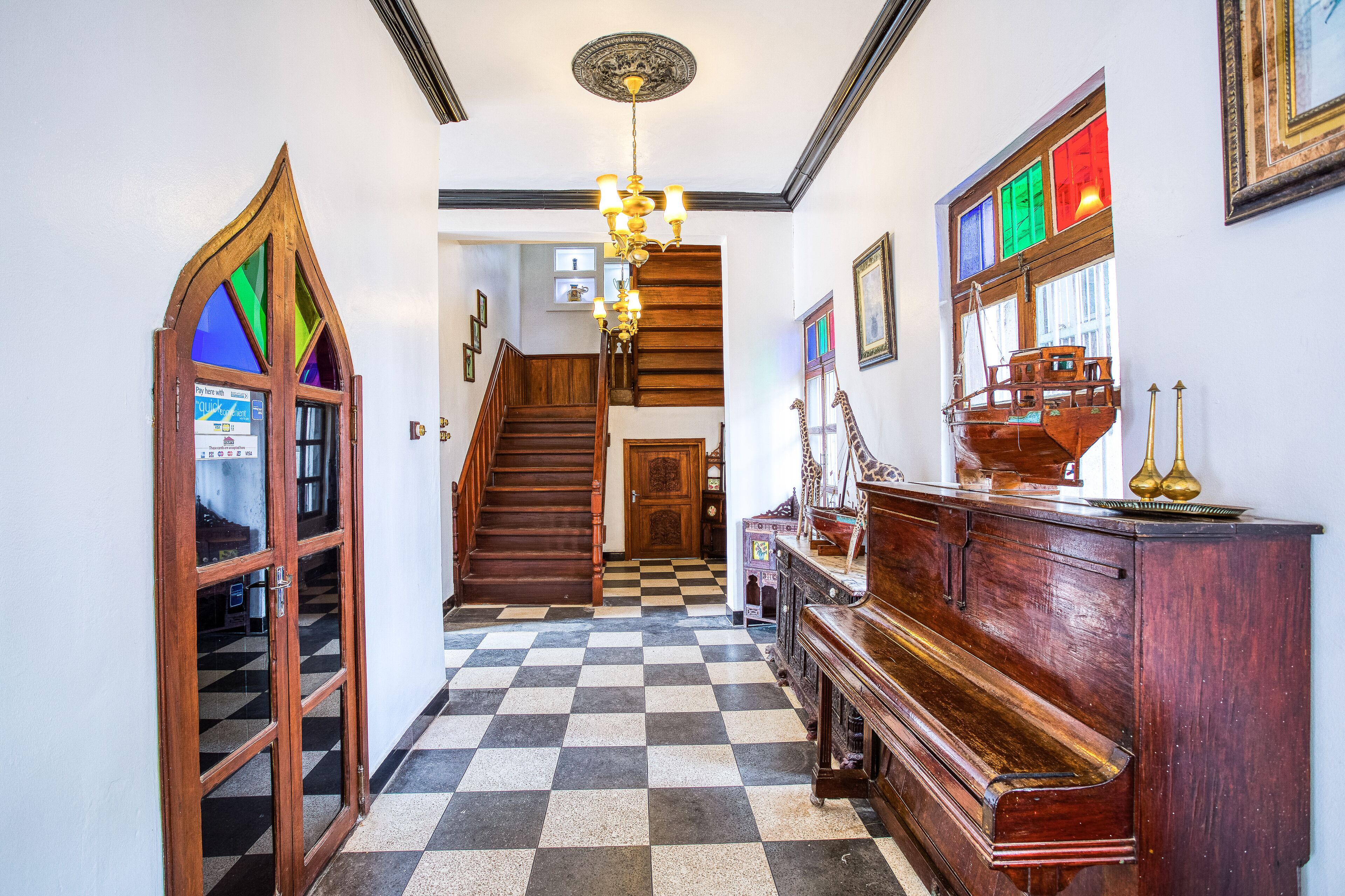 A checkered floor, wooden staircase, and a grand piano in a hallway.