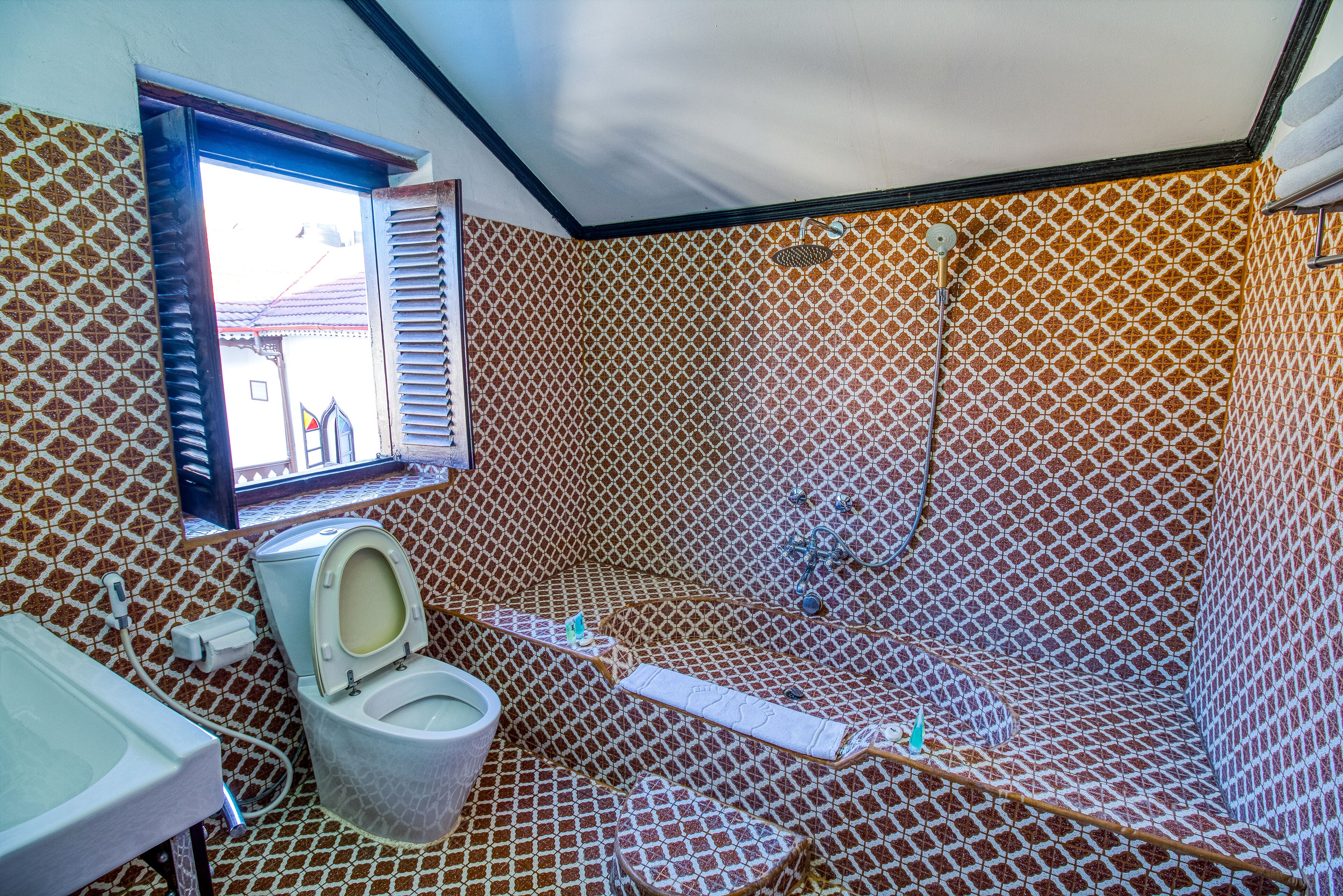 A bathroom with a patterned tile wall, a toilet, a shower, and a window with blinds.