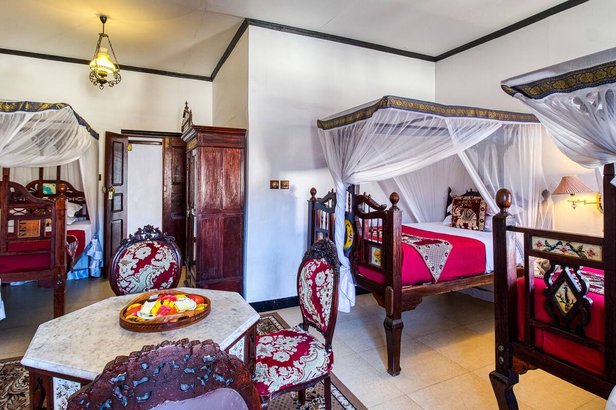 Une chambre traditionnelle avec un lit à baldaquin, une armoire en bois, une table sur laquelle se trouve un panier de fruits, et une chaise à motifs rouges et dorés.