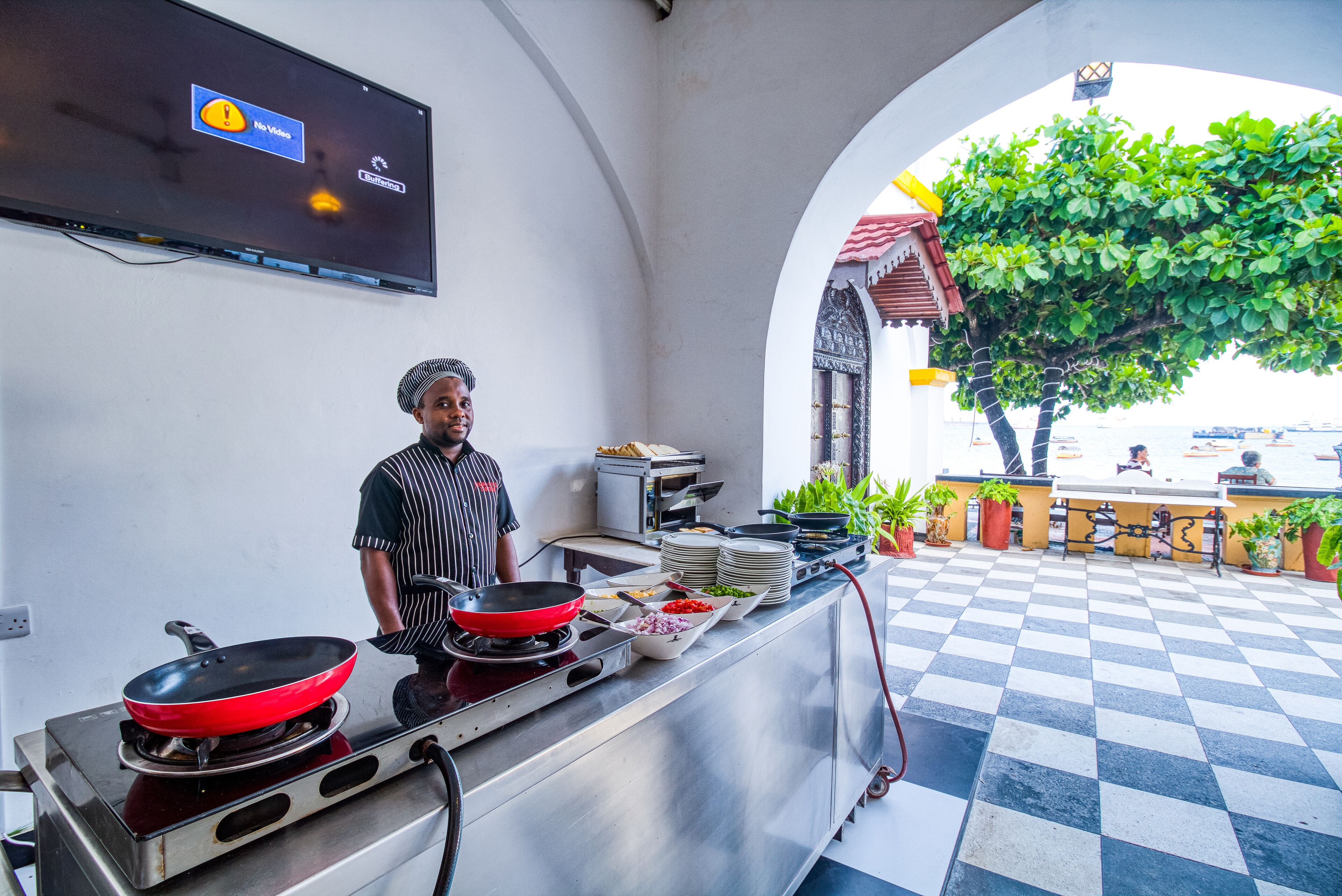 A chef in a kitchen with a TV displaying a message, a checkered floor, and potted plants.