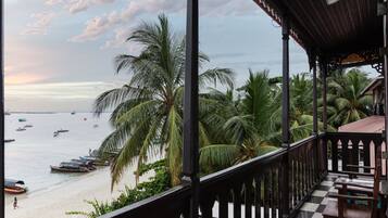 A balcony with a checkered floor, overlooking a beach with palm trees and boats.