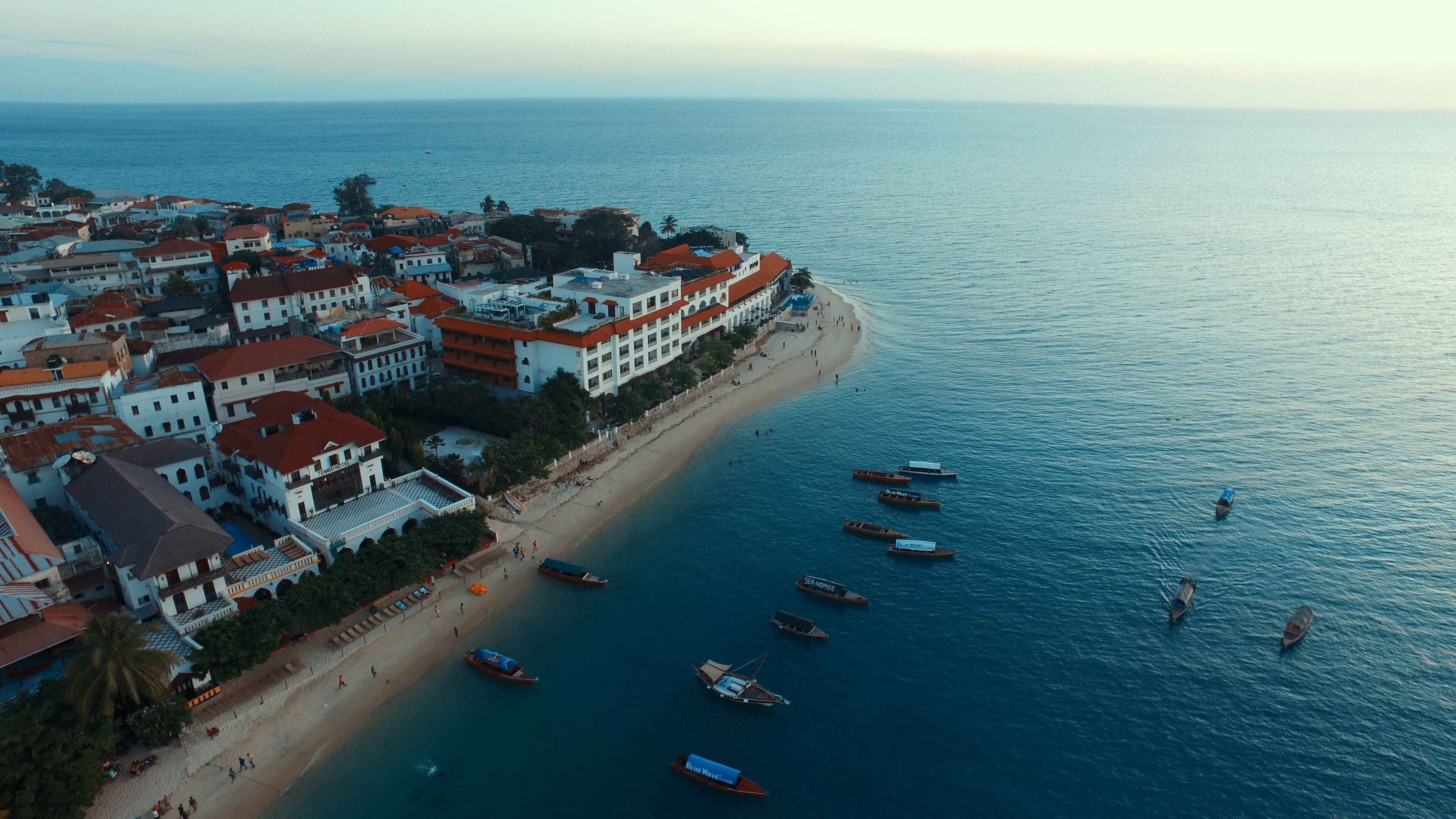 Vista aérea de un pueblo costero con playa, barcos y edificios con techos rojos.