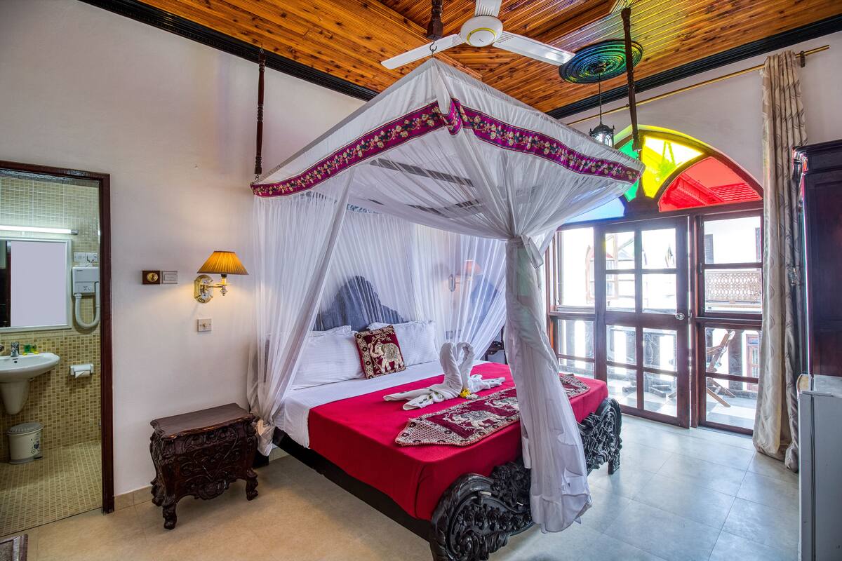 Une chambre à coucher avec un lit à baldaquin, une table de chevet, une commode, un ventilateur de plafond et une fenêtre avec des rideaux.