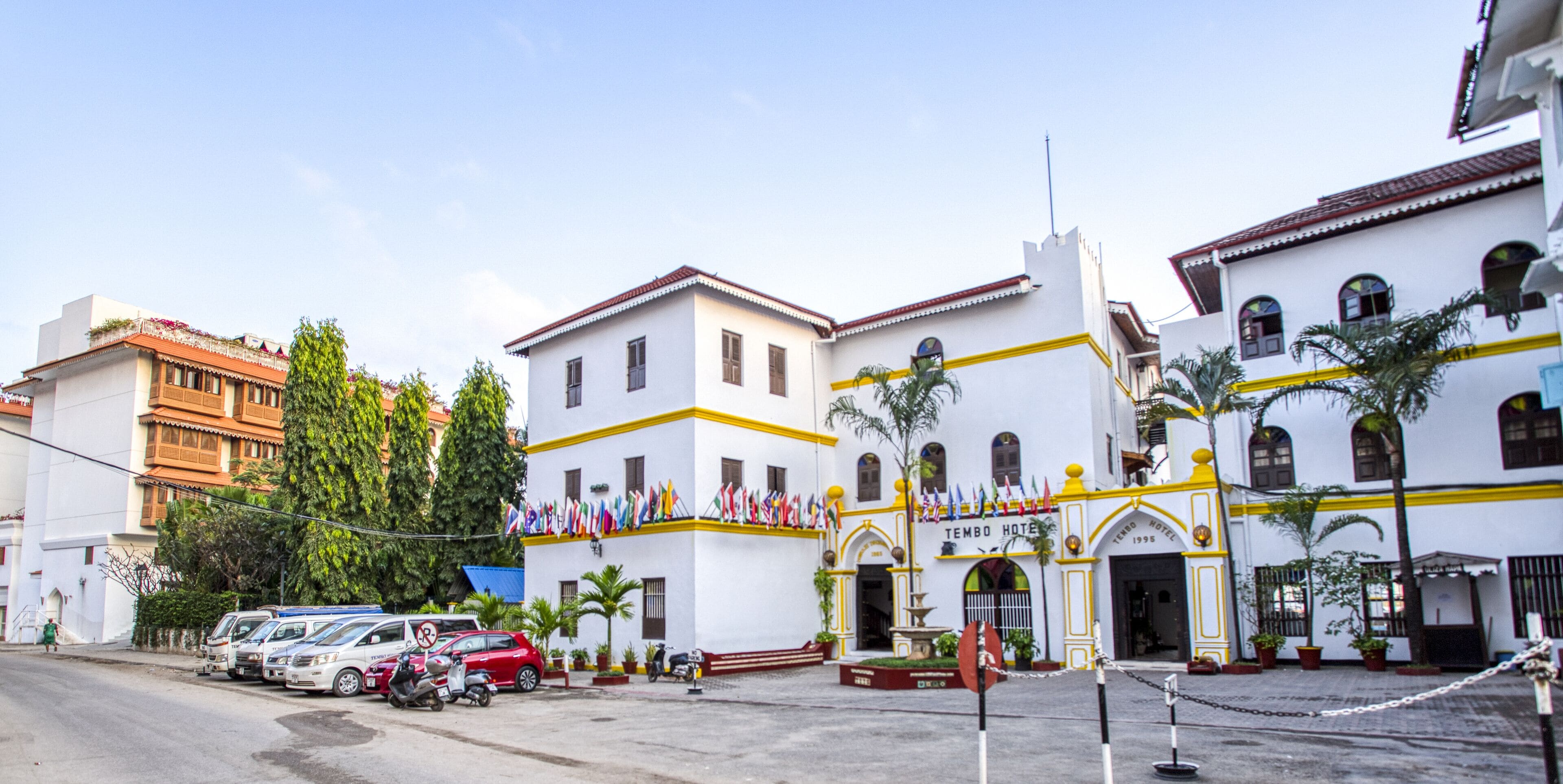 A hotel with a white facade, yellow trim, and multiple flags. There are parked vehicles and a fountain in front.