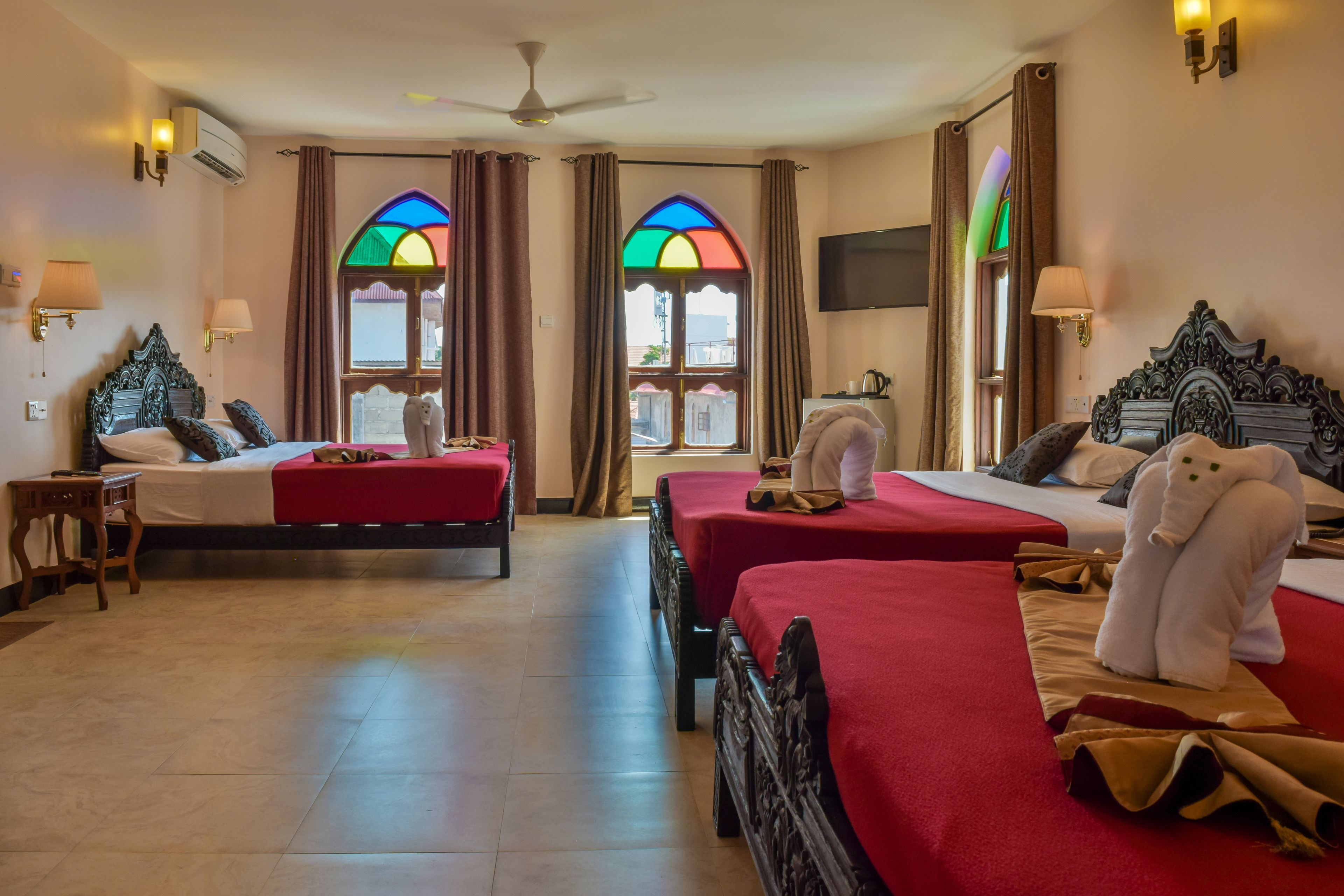 A hotel room with two beds, a ceiling fan, a TV, and a window with stained glass.