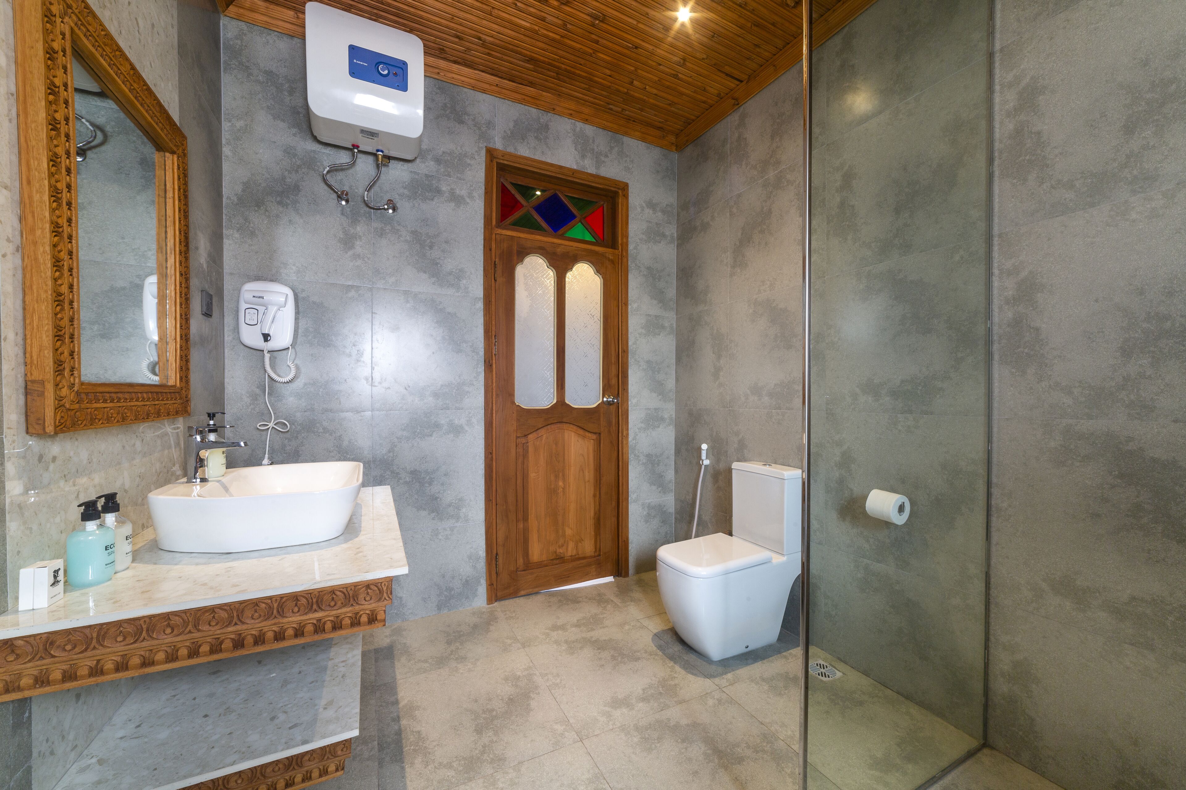 Une salle de bain avec un meuble-lavabo en bois, un lavabo blanc, une cabine de douche en verre et une porte en bois ornée de vitraux.