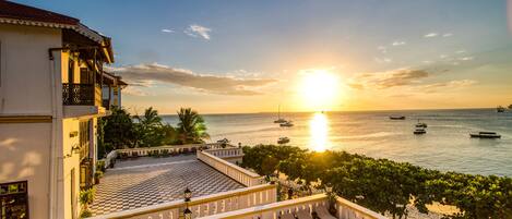 A terrace with checkered flooring, overlooking a body of water with boats and a sunset.