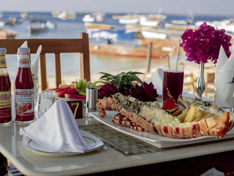 Una mesa dispuesta con un langostino, condimentos, una bebida y una flor, sobre una playa con barcos al fondo.