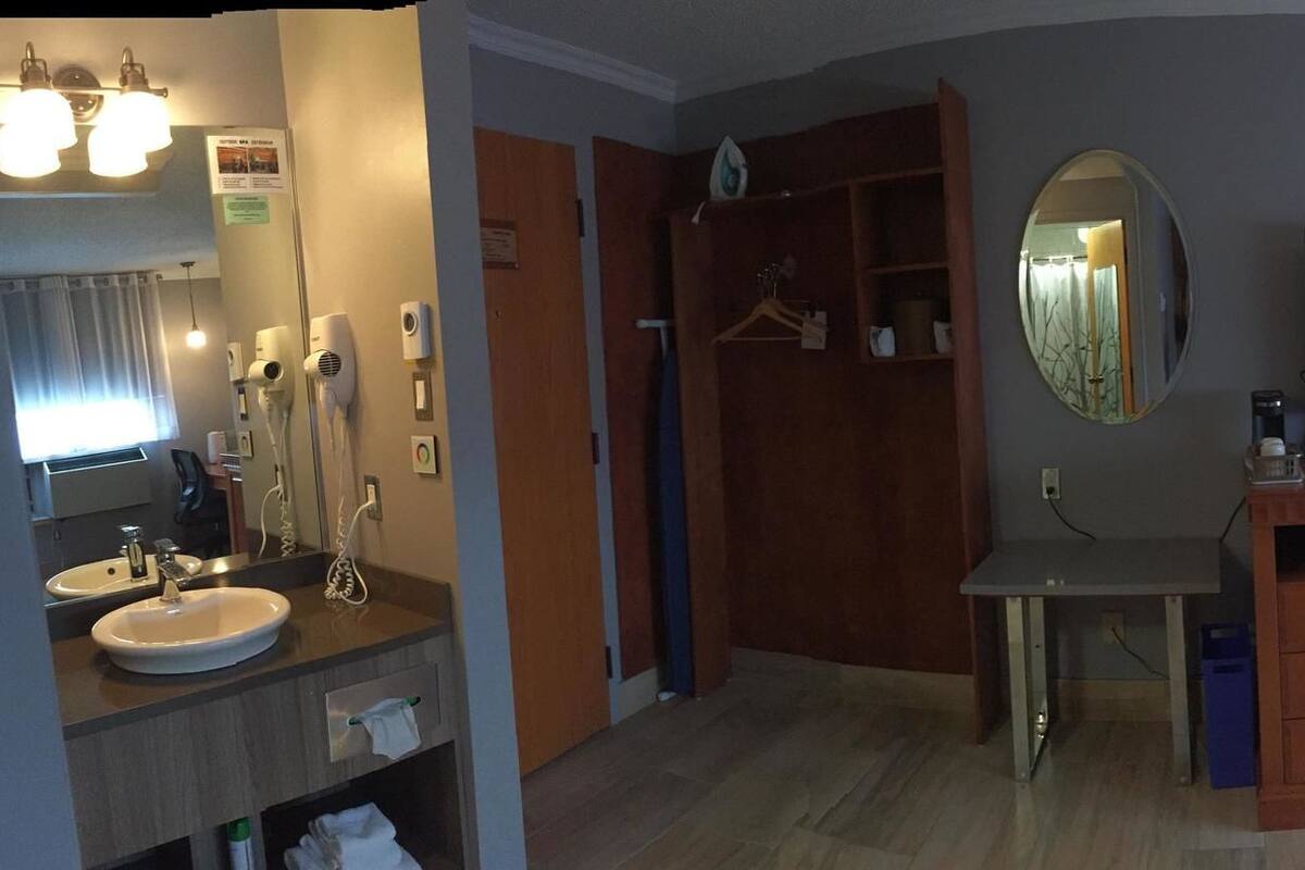A hotel room with a bed, a desk with a chair, a TV, a window with curtains, a mirror, and a bathroom with a sink and mirror.