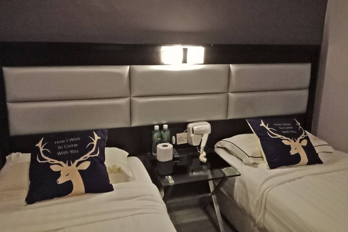 Habitación de hotel con dos camas, cada una con un almohadón que presenta un ciervo y el texto "How I Wish To Come With You". Una mesa de vidrio sostiene una botella, un dispensador de pañuelos y un cargador de teléfono.