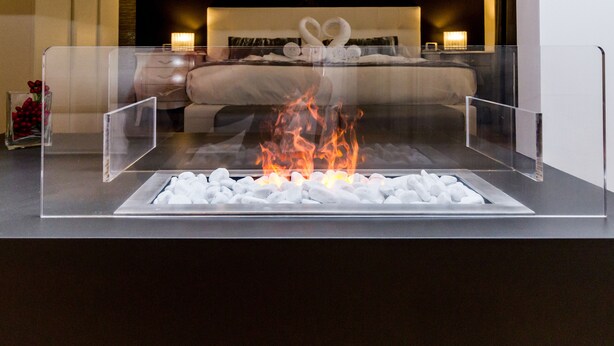 A modern hotel room with a glass-enclosed fireplace, a neatly made bed, and a nightstand with a lamp. - MyApART Suite (Rome)