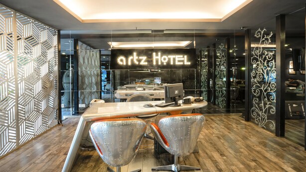 Een moderne hotel lobby met een receptiebalie, twee metalen stoelen en een decoratieve muur met geometrische patronen.