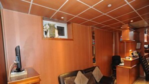 A room with a wooden panel wall, a brown leather sofa with cushions, a desk with a computer, and a small window with a decorative item. - Sully Hôtel (Paris)