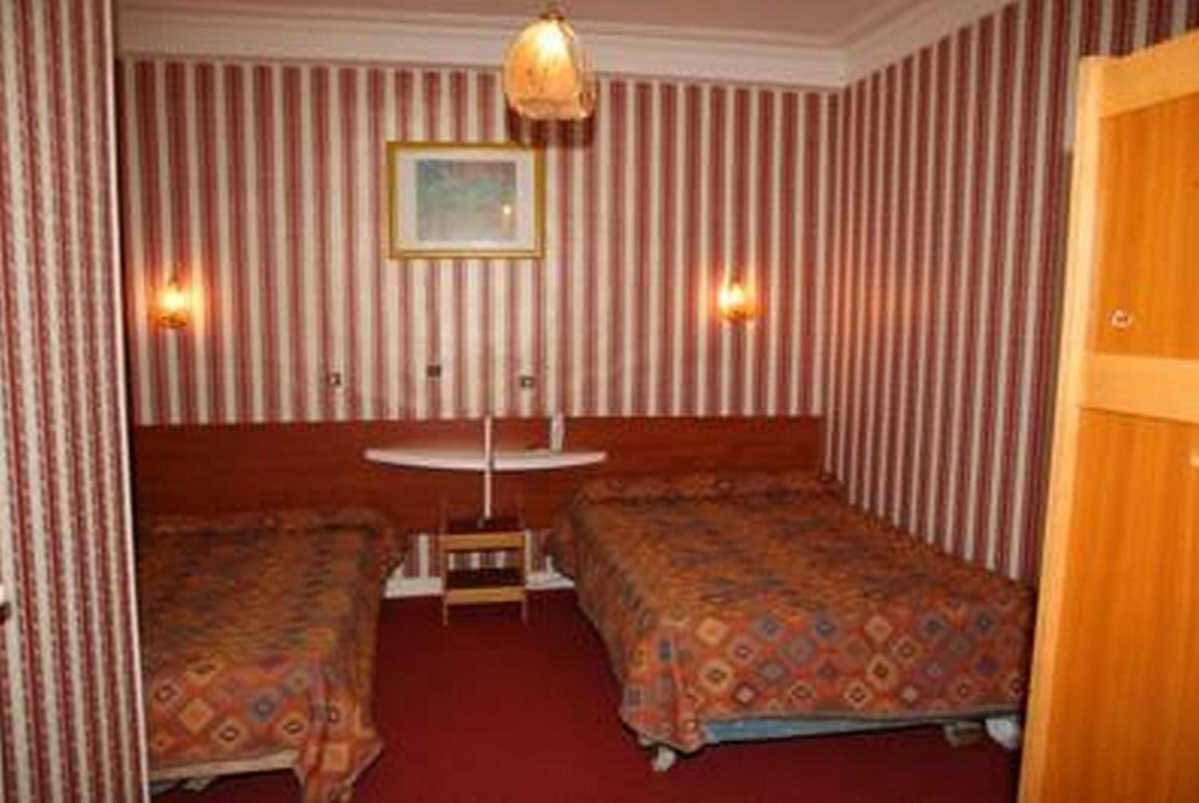 Ein Zimmer mit Streifenmuster-Tapete, zwei Betten mit geblümten Tagesdecken, ein runder Tisch und zwei Wandlampen.