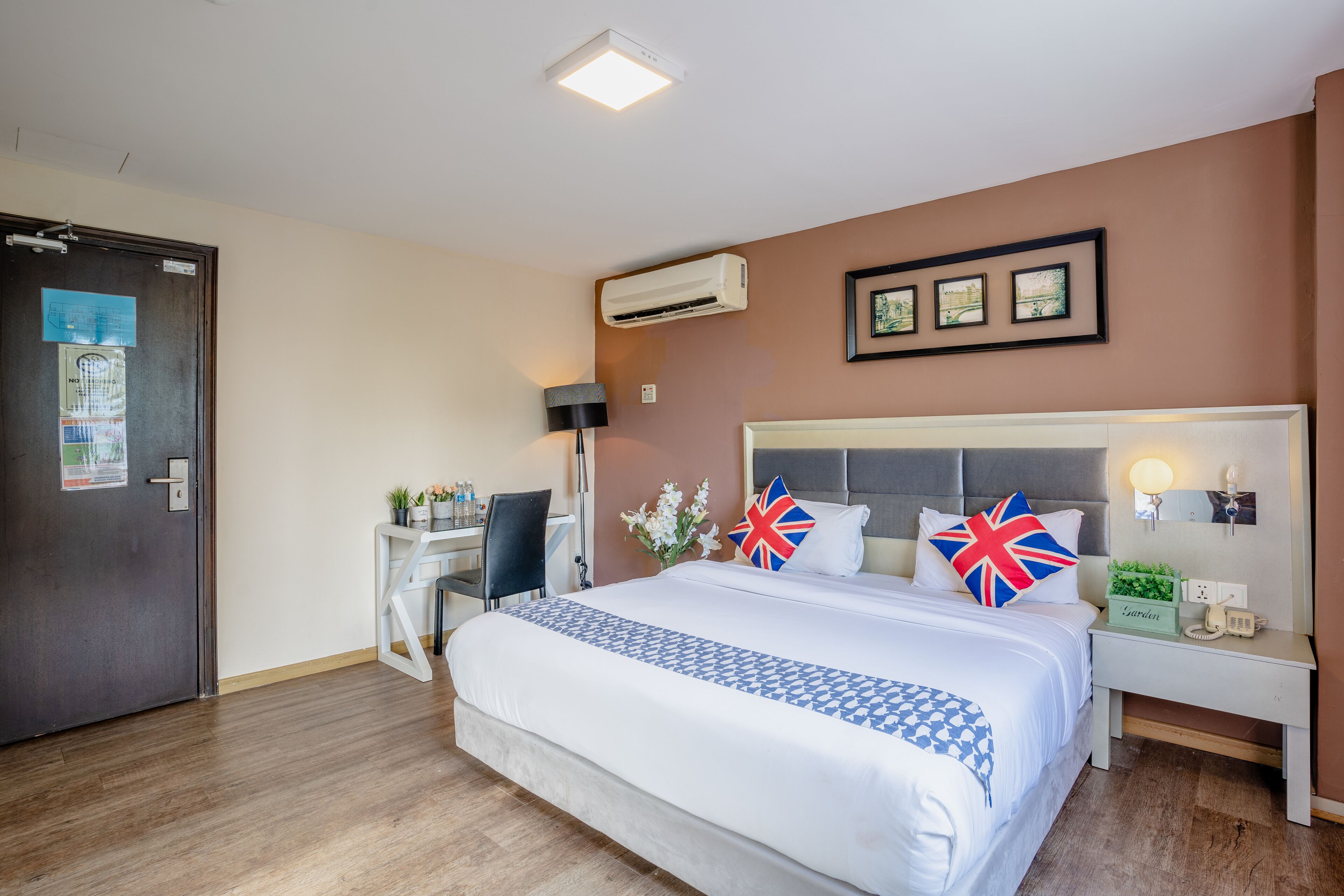 A hotel room with a large bed, a desk, a chair, an air conditioning unit, a lamp, a nightstand, and a wall-mounted phone.