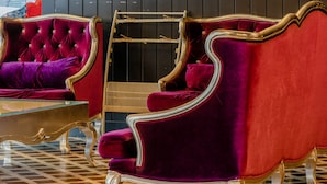 Une chambre luxueuse, ornée d’un lustre, comprenant deux fauteuils en velours rouge, un téléviseur fixé au mur et une pancarte indiquant « Coin journaux ».