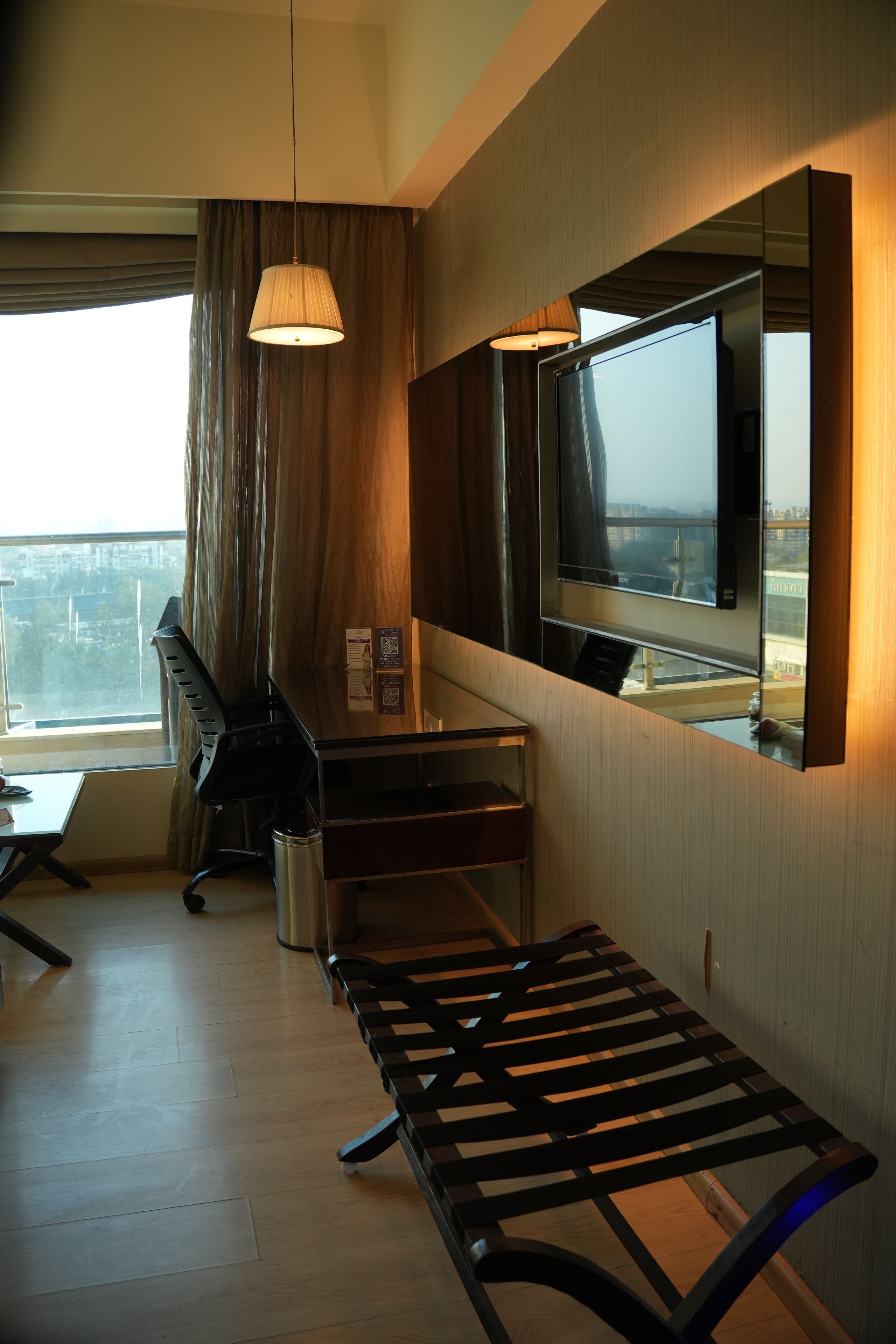A hotel room with a large window, a desk, a chair, a bench, a TV, and a hanging light fixture.