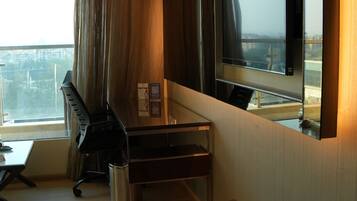 A hotel room with a large window, a desk, a chair, a bench, a TV, and a hanging light fixture.