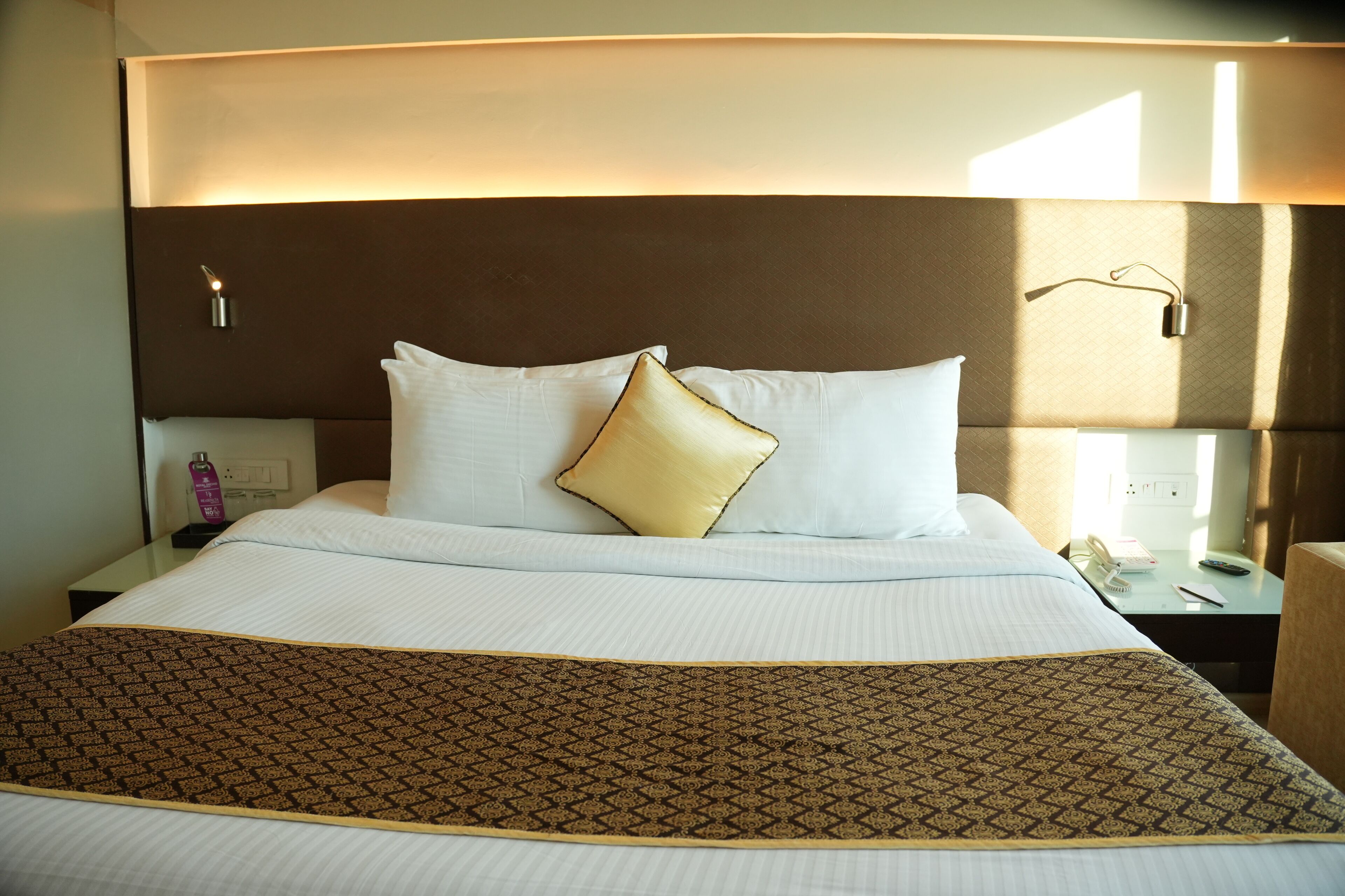 A neatly made bed with white linens and a patterned throw, a bedside table with a lamp, and a nightstand with a phone and a bottle.