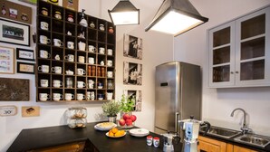 Cozinha com bancada preta, geladeira de aço inoxidável e armários de madeira. Há vários copos e objetos decorativos nas prateleiras, e uma planta sobre a bancada.