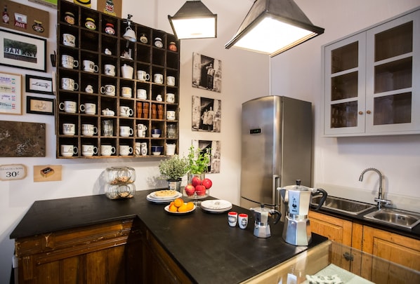 A kitchen with a black countertop, stainless steel refrigerator, and wooden cabinets. There are various mugs and decorative items on shelves, and a plant on the counter.