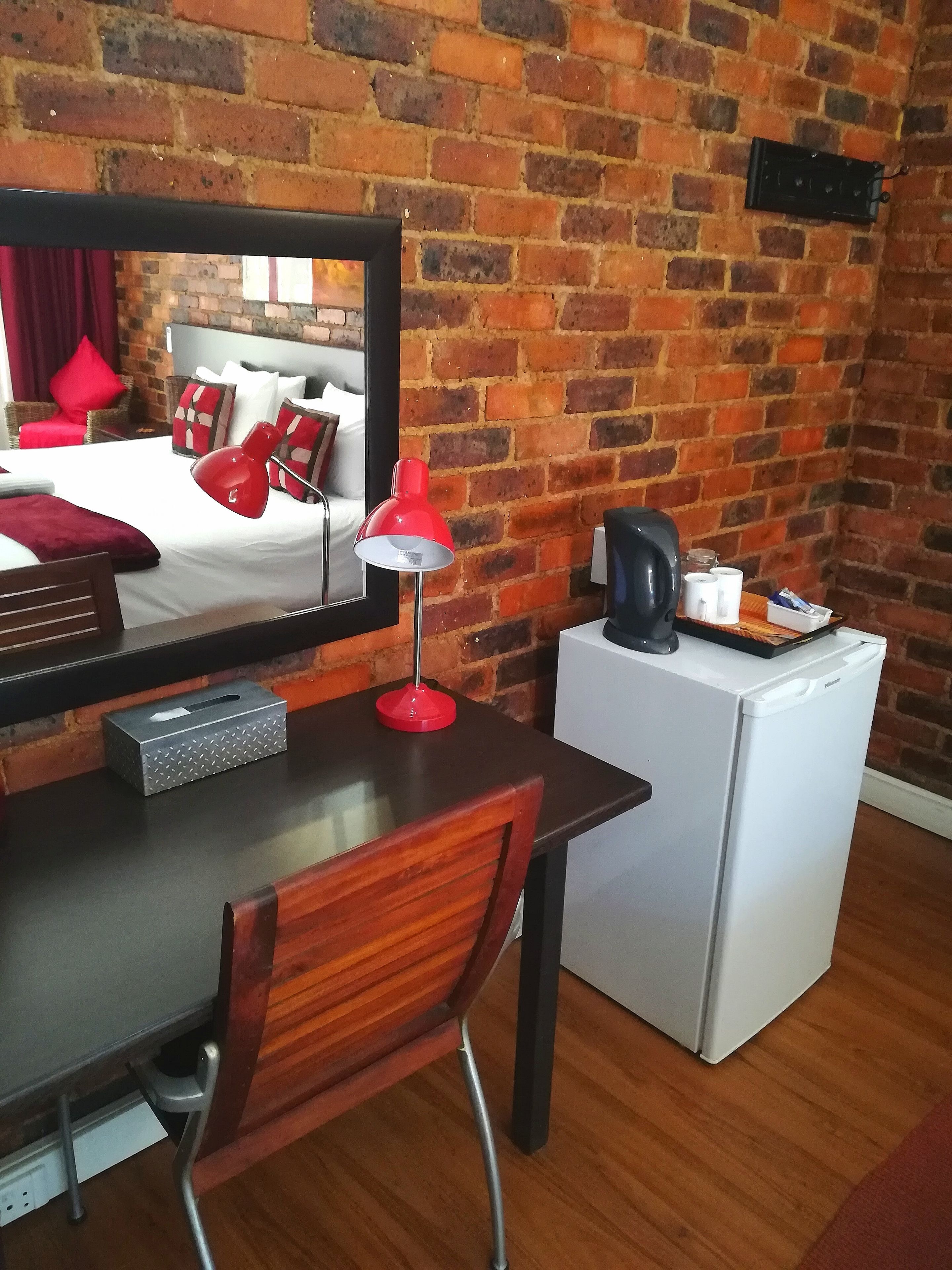 A hotel room with a brick wall, a bed with red bedding, a desk with a red lamp, a small refrigerator, and a mirror reflecting the room.