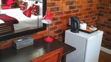 A hotel room with a brick wall, a bed with red bedding, a desk with a red lamp, a small refrigerator, and a mirror reflecting the room.