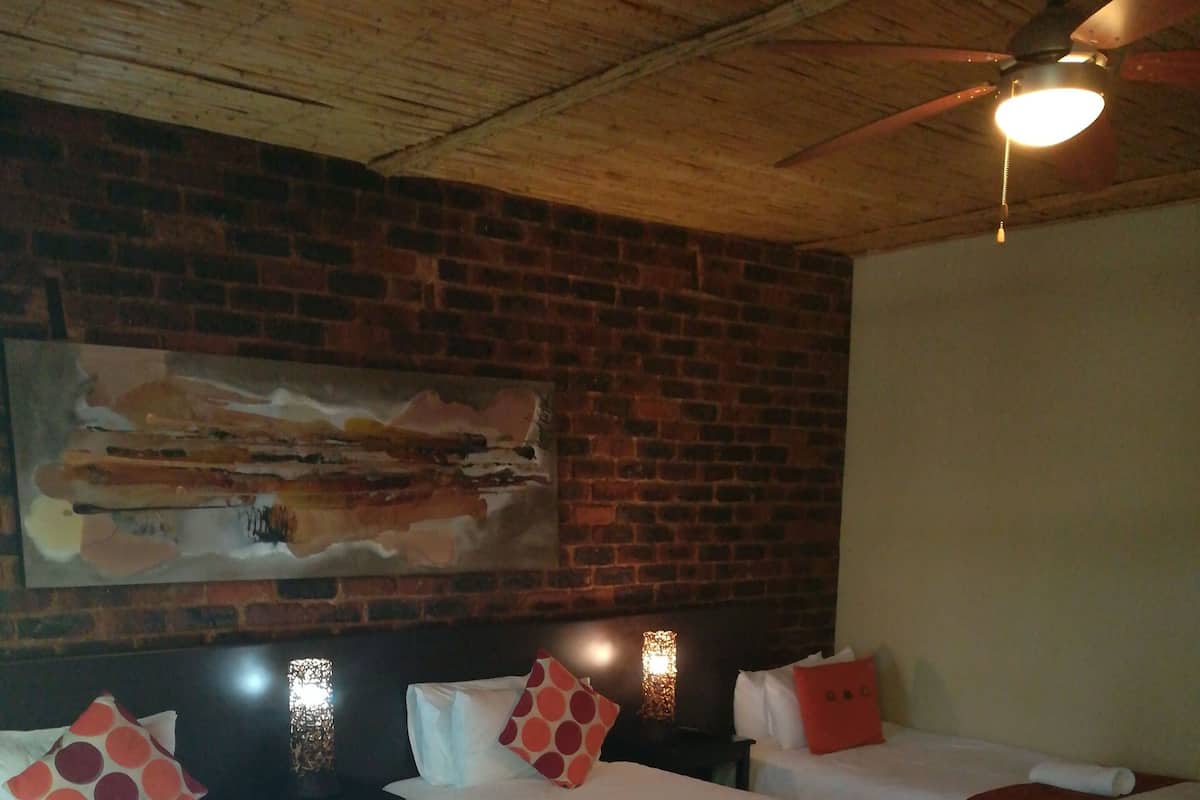 Ein Hotelzimmer mit vier Einzelbetten, jedes mit einer roten und weißen Decke, weißen Kissen und einer roten Tagesdecke. Das Zimmer hat eine Backsteinwand und eine Holzbalkendecke mit Deckenventilator.